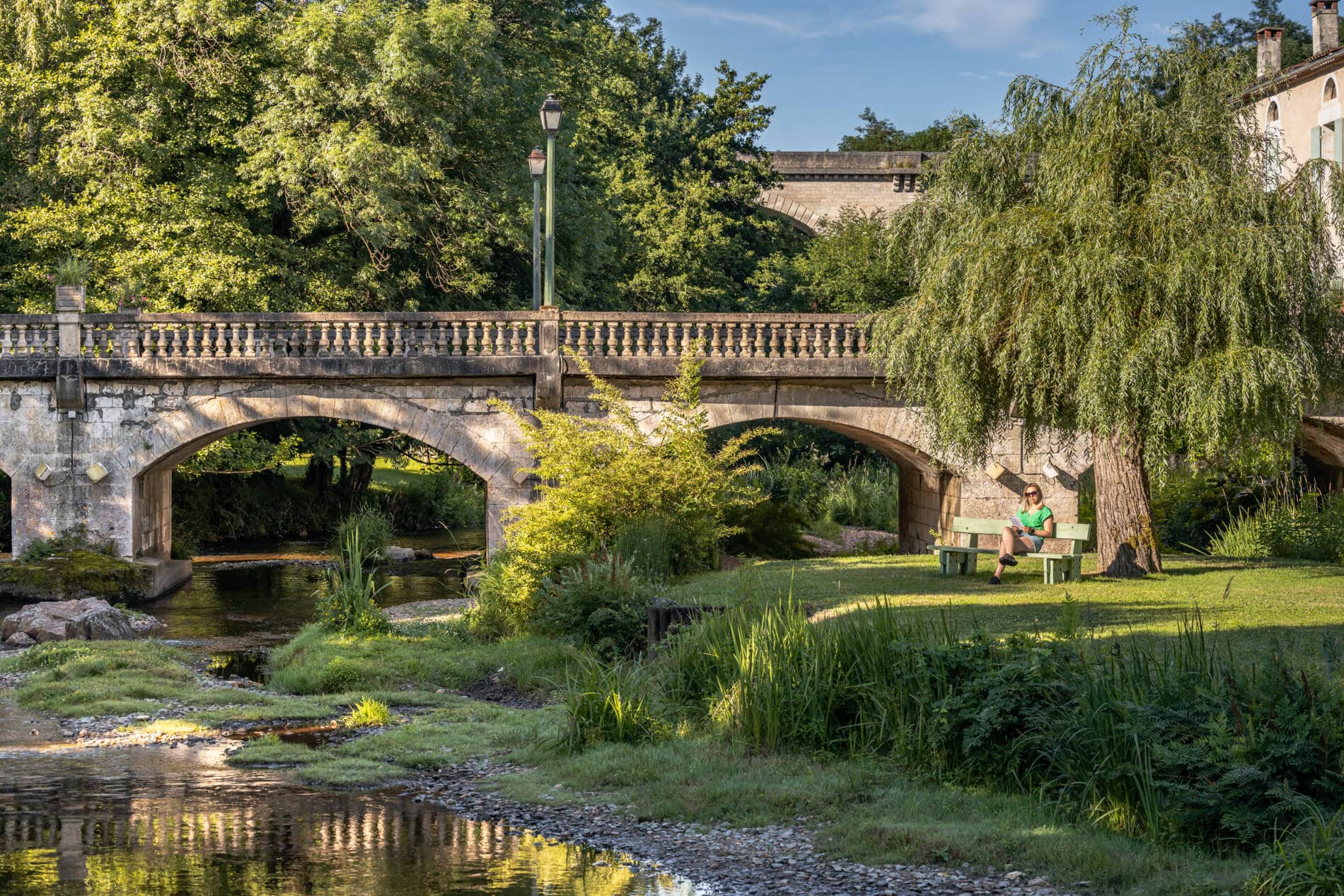 Un pont au-dessus d'une rivière. Au pied du pont un saule et un banc. Une jeune femme, assise sur le banc, est en train de lire.